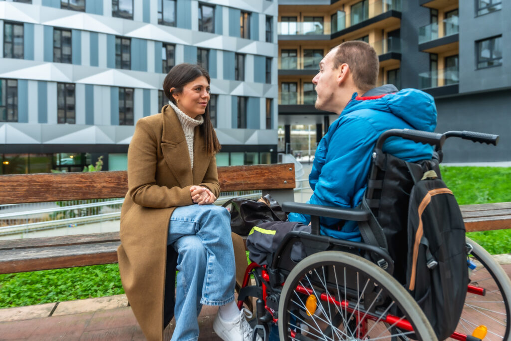 Assistenzkraft und junger Mann im Rollstuhl im Gespräch auf einer Parkbank in der Stadt – Selbstbestimmung und Teilhabe mit 24h Assistance Care Hamburg
